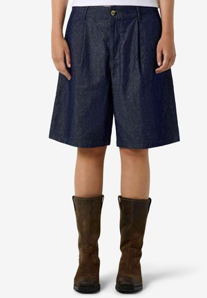 Person iført mørkeblå vidde shorts og brun læderstøvler til midt på læggen står mod en hvid baggrund.