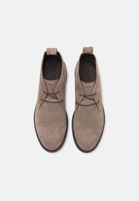 Botas chukka en gamuza de color marrón claro con un acabado suave, con cordones de cuero y punta redondeada. El logo de la marca está embosado en la plantilla.