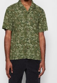 Maglietta verde a maniche corte con motivo a foglie, caratterizzata da un design con bottoni sul davanti e una texture sottile. Ideale per un abbigliamento casual.