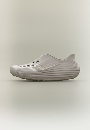 Πλευρική όψη ενός ανοιχτό μπεζ παπουτσιού slip-on με οπές αερισμού, υφασμένες ραβδώσεις και ασημένιο λογότυπο Nike σε απλό φόντο.