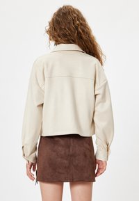 Chaqueta beige recortada con un corte relajado, que presenta un cuello clásico y mangas largas. Combinada con una mini falda de ante marrón.