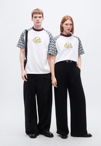 Λευκά T-shirts με μανίκια με σχέδια, που φέρουν κίτρινο κυκλικό λογότυπο. Φοριούνται με φαρδιά μαύρα παντελόνια. Μινιμαλιστικός σχεδιασμός και αντιθετικά χρώματα.