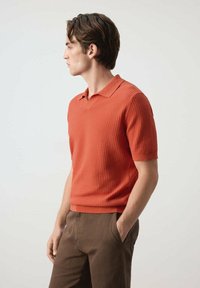 Jeune homme en polo orange texturé à manches courtes et pantalon marron, une main dans la poche, face à gauche sur fond uni.
