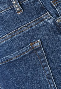 Vaqueros de mezclilla con un lavado azul oscuro, con un ligero desgastado en la parte delantera, costuras de color cobre y herrajes metálicos en la cintura.