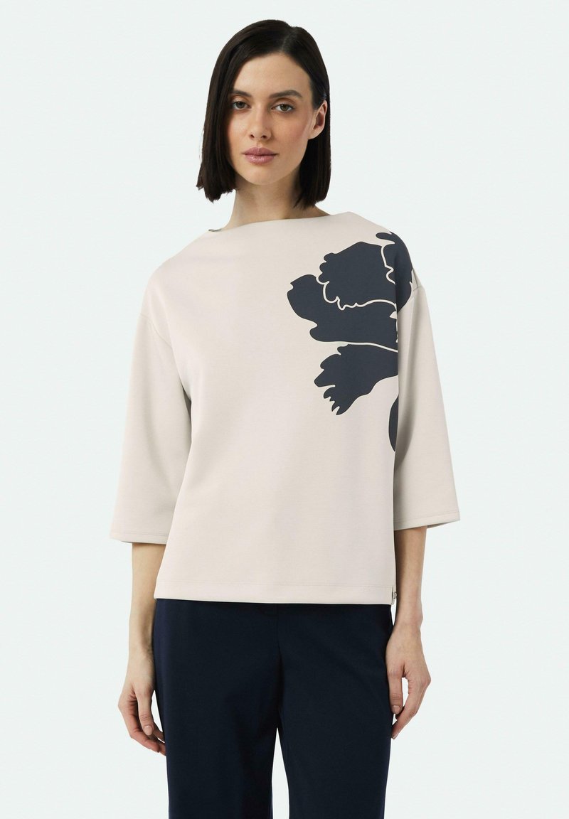 Light beige three-quarter sleeve top with a dark floral design on the left side, soft texture, and boxy silhouette.