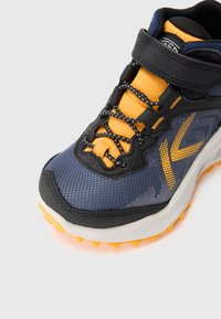 Sport shoe featuring a dark blue mesh upper, bright orange accents, black rubber trim, and textured sole with orange spikes. Adjustable strap included.