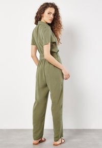 Olivgrüner Jumpsuit mit kurzen Ärmeln, Kragen und tailliertem Gürtel. Aus strukturiertem Stoff gefertigt, mit geradem Bein und schlichten Sandalen.