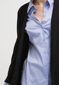 Blazer negro superpuesto sobre una camisa de botón azul claro. La camisa tiene un cuello clásico y pequeños botones, con una textura suave.