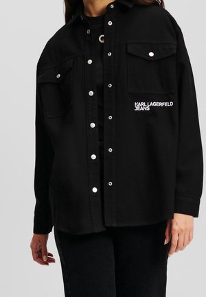 Person wearing a black button-up jacket with silver snaps, chest pockets, and "KARL LAGERFELD JEANS" embroidered on the front.