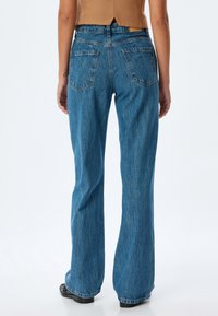 Vaqueros de mezclilla azul con forma de campana y cintura alta, bolsillos traseros visibles y un ligero desgaste; textura suave con un sutil brillo.