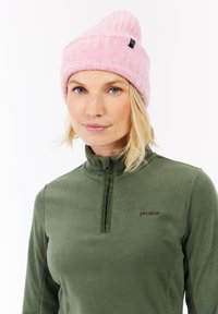 Kvinde med blondt hår iført en pink strikket hue og en olivengrøn fleece-overdel med kvart lynlås, som står vendt fremad med en neutral udtryk.