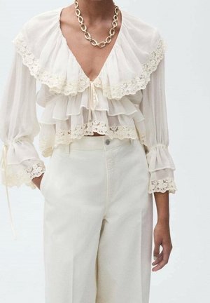 Vrouw draagt witte gelaagde kanten blouse met grote kraag, 3/4 mouwen, gecombineerd met witte hoge taille broek en een dikke ketting halsketting.