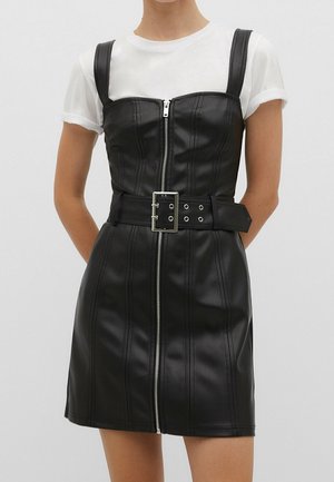 Vrouw draagt een zwarte, nauwsluitende jurk van nep leer met ritssluiting aan de voorkant, brede riem met zilveren gesp, over een wit T-shirt met korte mouwen.