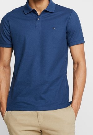 Homme portant un polo bleu ajusté à manches courtes avec un petit logo blanc sur la poitrine et un pantalon beige.