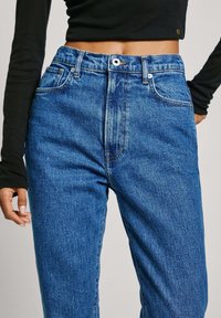 Sinisenväriset korkeat farkut, joissa haalistunut pesu, etutaskut, vetoketjullinen sivusauma ja metalliset napit. Kankaan pinta on teksturoitu.