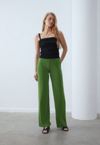 Haut noir froncé avec des fines bretelles associé à un pantalon vert taille haute et à jambes larges. Les deux vêtements ont des textures lisses. Sandales noires à talons.