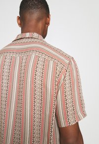 Kurzarmpullover aus beigem Stoff mit roten und braunen geometrischen Mustern. Auffällige gewebte Textur und Detail am Kragen.