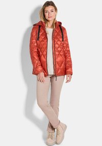 Gesteppten orange Jacke mit Kapuze, schwarzen Akzenten und Reißverschluss; kombiniert mit einem cremfarbenen Pullover, beigen Hosen und hellbeigen Schuhen.