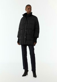 Czarna kurtka puchowa o pudełkowym kształcie, zapinana na napy, z wysokim kołnierzem i ściągaczami przy mankietach. W zestawie z ciemnymi dżinsami i spiczastymi botkami za kostkę.