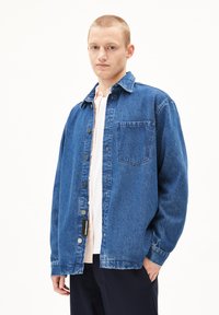 Jeansjack in medium blauw, met een knoopsluiting aan de voorkant, klassieke kraag en een enkele borstzak. De stof heeft een zachte, gestructureerde afwerking.