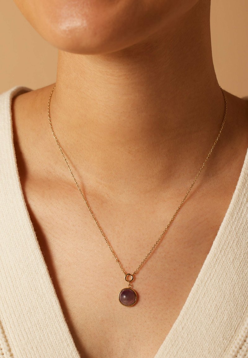 Collier en chaîne dorée avec un pendentif en gemme ronde de couleur violette. La chaîne a une texture délicate, mettant en valeur la pierre lisse.