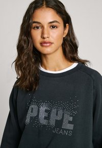 Sudadera negra con cuello redondo, que presenta un logo puntiado "PEPE JEANS" en plata. La tela parece suave y tiene un corte holgado.