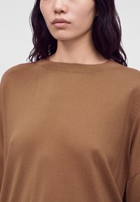 Femme aux longs cheveux foncés portant un pull en maille marron clair sur un fond uni.