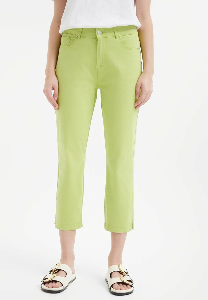 Zielone bawełniane spodnie 3/4 o dopasowanym kroju, z kieszeniami z przodu i małymi rozcięciami po bokach u dołu. Połączone z białymi sandałami z złotymi akcentami.