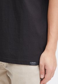Black cotton t-shirt with a smooth texture, showing a side view with a label reading "11 PROJECT" at the hem, paired with beige pants.