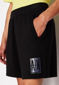 Pantaloni shorts neri realizzati in tessuto morbido con vita elasticizzata, dotati di tasche laterali e un logo riflettente Armani Exchange sulla coscia sinistra.