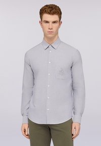 Camisa de manga larga gris claro con cuello, cierre frontal de botones y un bolsillo en el pecho que presenta un logotipo bordado. Textura suave.