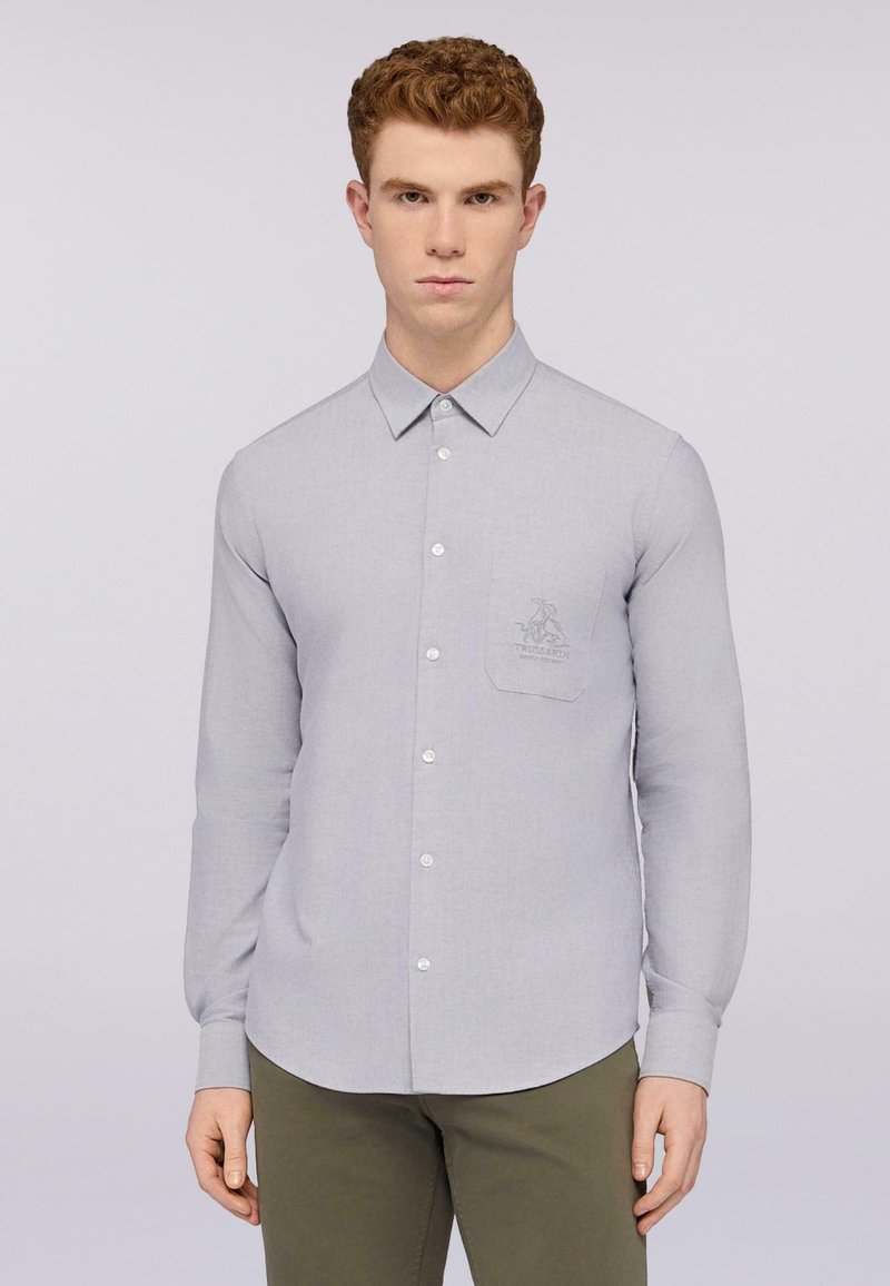 Camisa de manga larga gris claro con cuello, cierre frontal de botones y un bolsillo en el pecho que presenta un logotipo bordado. Textura suave.