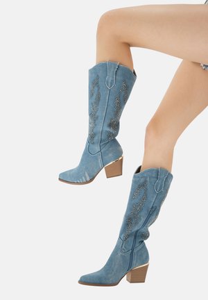 Botas de mezclilla azul hasta la rodilla con adornos decorativos de tachuelas plateadas, punteras puntiagudas y un tacón apilado. Presentan costuras de estilo western y Acabados elevados.