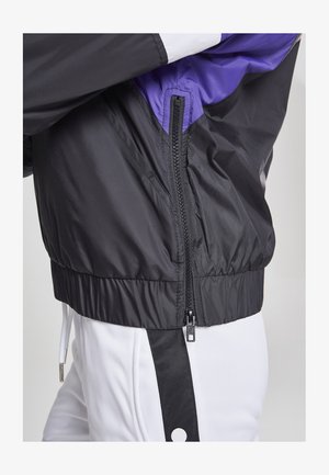 Črno vetrovko z vijoličnimi detajli, ki ima zadrgo, rebraste manšete in pas. Gladka tekstura tkanine, lahka zasnova.