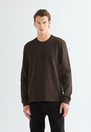 Jonge man met een effen donkerbruine lange mouw shirt en zwarte broek, staand tegen een egale lichte achtergrond.