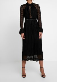 Robe en dentelle noire avec manches longues, col haut, texture plissée et ourlet festonné. Comprend une ceinture cintrée et des poignets accentués.