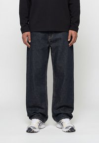 Άνδρας που φοράει φαρδιά σκούρα μπλε τζιν, λευκά και γκρι αθλητικά παπούτσια και μαύρο μακρυμάνικο πουκάμισο, στέκεται μπροστά σε απλό φόντο.