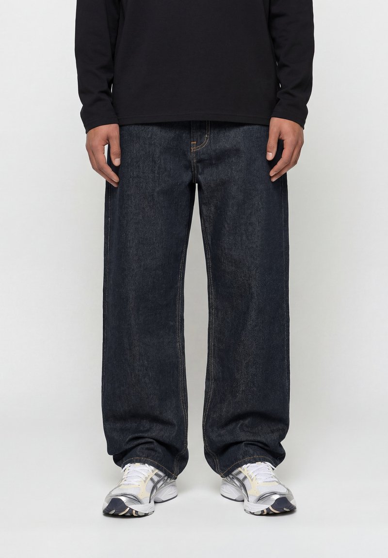 Άνδρας που φοράει φαρδιά σκούρα μπλε τζιν, λευκά και γκρι αθλητικά παπούτσια και μαύρο μακρυμάνικο πουκάμισο, στέκεται μπροστά σε απλό φόντο.