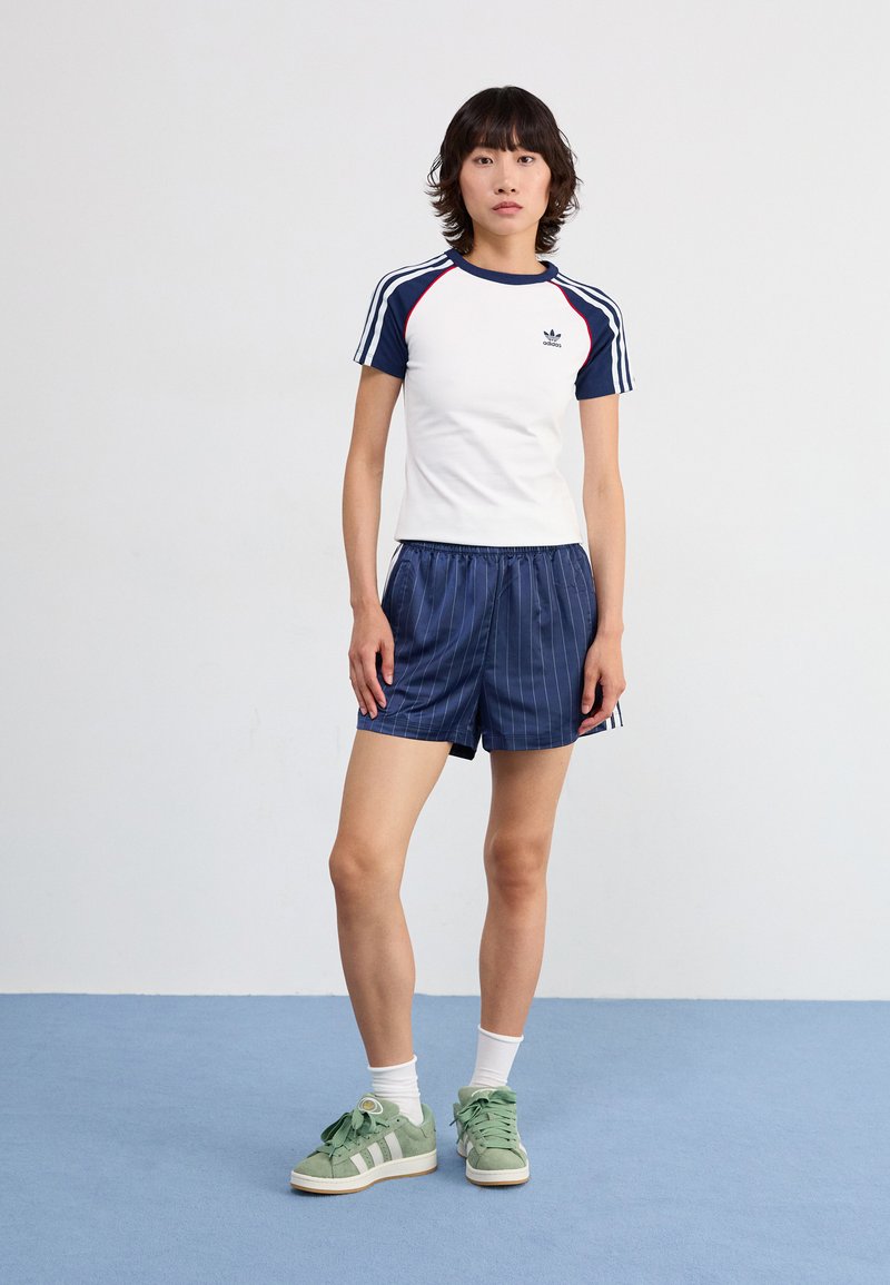 Camisa deportiva blanca con mangas azul marino, detalles de rayas y logo. Pantalones cortos azul marino a rayas. Zapatillas verdes con rayas y cordones blancos.