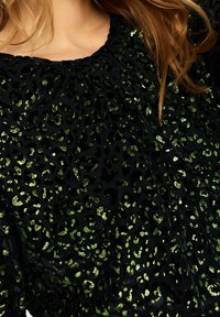 Haut noir avec des taches de léopard vert brillantes, texture douce, encolure froncée et manches amples. Couture centrale visible sur le tissu.