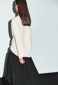 Blouse blanche à manches longues avec une coupe décontractée, associée à une jupe plissée noire. Le tissu lisse et la brillance subtile mettent en valeur la silhouette.