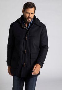 Schwarzer Wollmantel mit Toggle-Verschlüssen, großen Vordertaschen und Kapuze. Kombiniert mit einem blauen Hemd und Jeans, was einen lässigen Look zeigt.