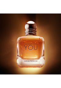 Bottiglia di profumo in vetro trasparente con liquido ambrato, tappo metallico rotondo e testo "Stronger With You Emporio Armani" sul davanti, sfondo dolcemente illuminato.