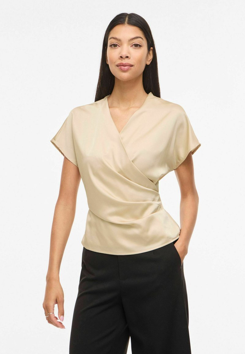 Žena s dlhými rovnými vlasmi, oblečená v béžovej zavinovacej blúzke s krátkym rukávom a čiernych nohaviciach, stojaca s jednou rukou v vrecku na bielom pozadí.