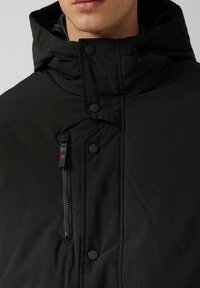 Schwarze Daunenjacke mit hohem Kragen, ausgestattet mit Druckknöpfen und einer Reißverschlusstasche. Das strukturierte Material sorgt für eine matte Oberfläche.