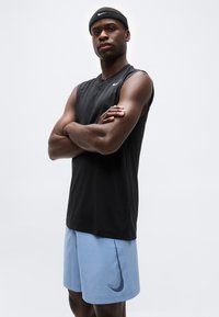 Αθλητικός άνδρας που φοράει μαύρο αμάνικο μπλουζάκι Nike, ανοιχτό μπλε σορτς Nike και μαύρη κορδέλα Nike στο κεφάλι, στέκεται με τα χέρια σταυρωμένα μπροστά σε λευκό φόντο.