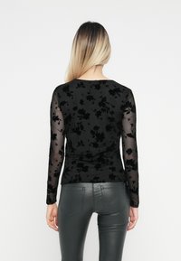Sort langærmet top med blomster-spidsoverdel og gennemsigtige ærmer. Tætsiddende design, glat tekstur. Sammen med skinnende mørke leggings.