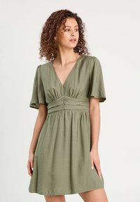 Robe vert clair avec de courtes manches papillon, un décolleté en V et des détails froncés à la taille. Tissu doux avec une jupe fluide.