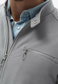 Grijze zip-up jack met gestructureerde stof, zilverkleurige hardware en een borstzak. Gelaagd over een lichtblauw overhemd met kraag.