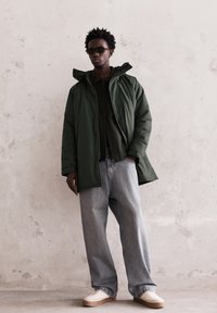 Bărbat îmbrăcat într-o parka verde închis, geacă neagră, blugi largi bleumarin deschis, adidași albi și ochelari de soare negri, pe fondul unei pereți texturate de culoare deschisă.